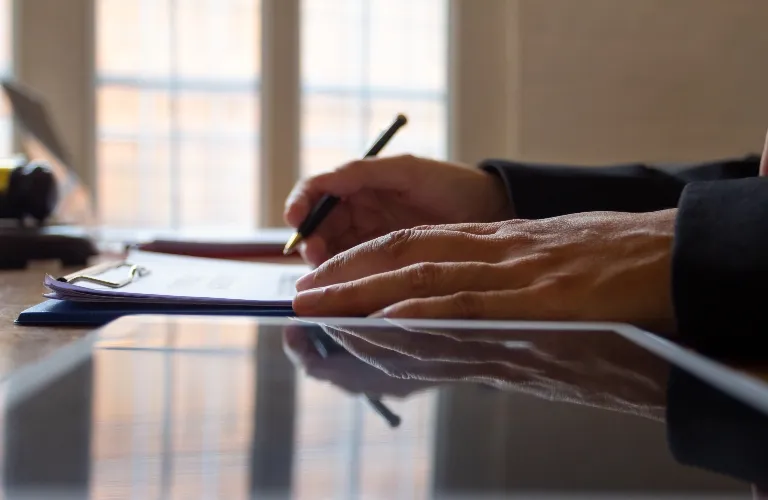 Close-up of hands writing on a clipboard with a pen beside a reflective tablet on a desk.