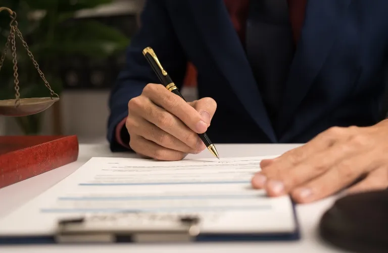 Person in dark suit signing document on clipboard with scales of justice nearby.
