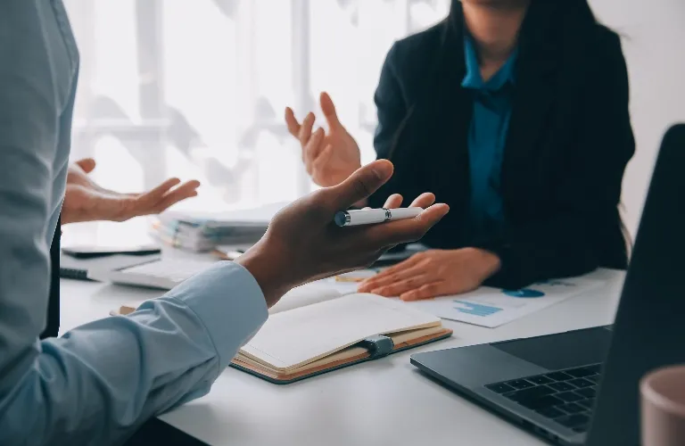Two professionals engaging in discussion at a desk with a laptop, notebook, and documents.