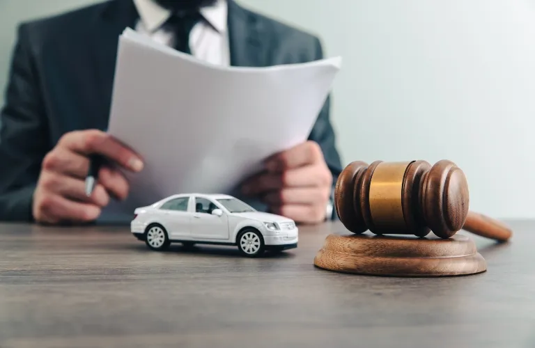 Wooden judge's gavel and white toy car on desk with person in suit holding documents.