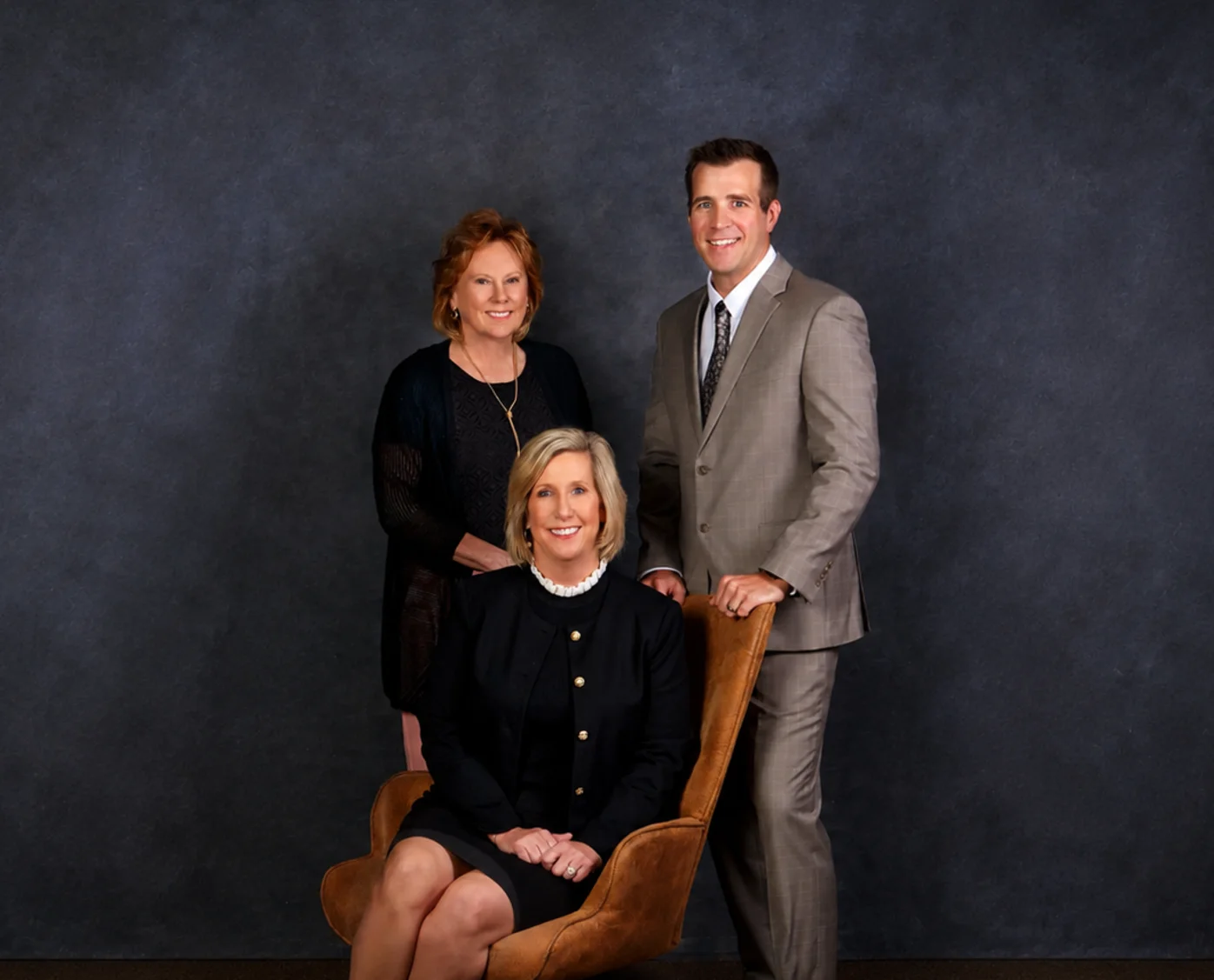 Professional portrait of two women and one man in formal attire against a dark gray backdrop.